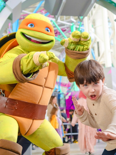Child posing with Teenage Mutant Ninja Turtle character at an indoor amusement park.