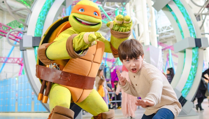 Child posing with Teenage Mutant Ninja Turtle character at an indoor amusement park.