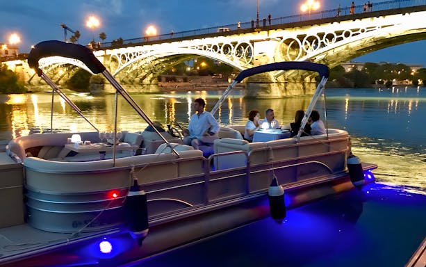 Tourists on a boat enjoying Guadalquivir River cruise with tapas and drinks near illuminated bridge.