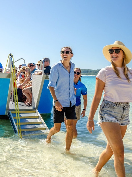 Tourists disembarking a boat at Whitehaven Beach, Airlie Beach, with clear blue water and lush hills.