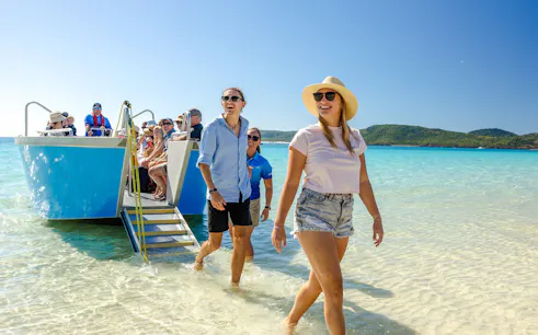 Tourists disembarking a boat at Whitehaven Beach, Airlie Beach, with clear blue water and lush hills.