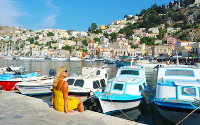 Woman sitting by boats at Symi Island harbor, Rhodes, with colorful hillside houses in the background.