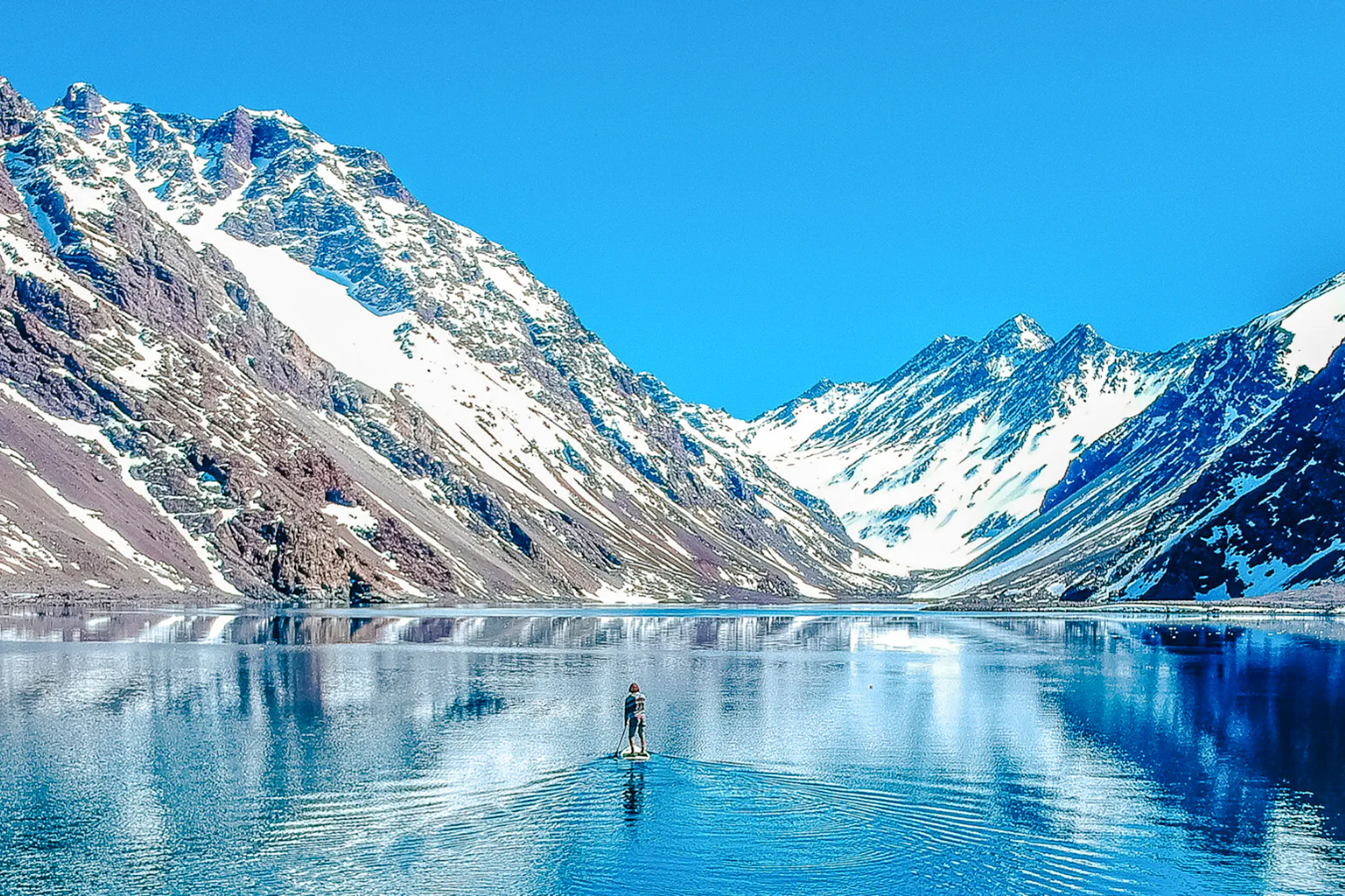 Person paddleboarding on Laguna del Inca with snow-capped Andes mountains, Portillo, Chile.