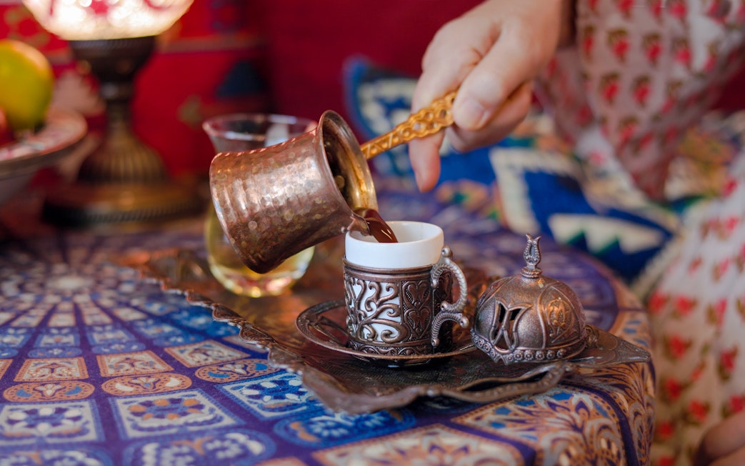 Pouring Turkish coffee from a copper cezve into an ornate cup on a patterned table.