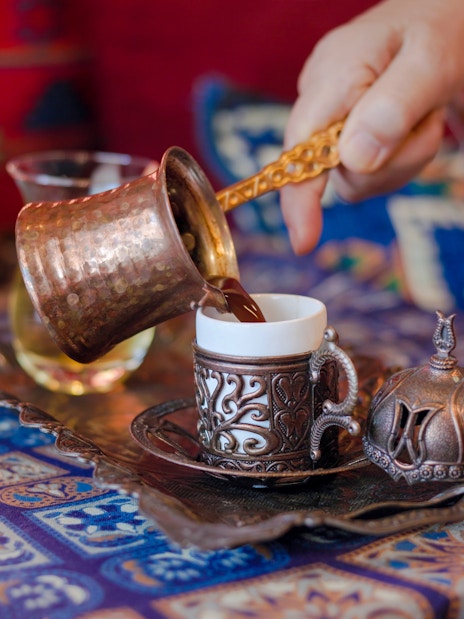 Pouring Turkish coffee from a copper cezve into an ornate cup on a patterned table.