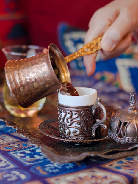 Pouring Turkish coffee from a copper cezve into an ornate cup on a patterned table.
