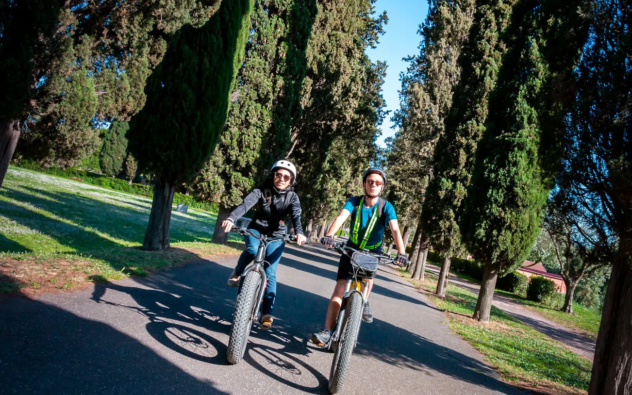 Cyclists on the Appian Way surrounded by trees in the Roman countryside.