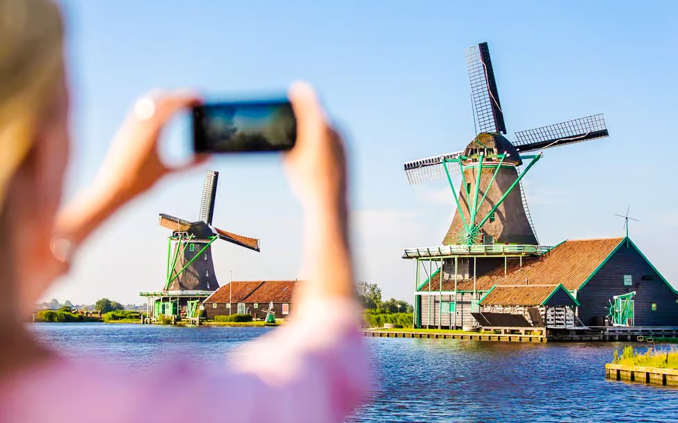 Windmills by the water in Volendam, Netherlands, with a person taking a photo.