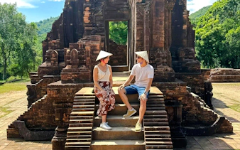 Visitors sitting on steps at Mỹ Sơn Sanctuary, Vietnam, with ancient temple ruins in the background.
