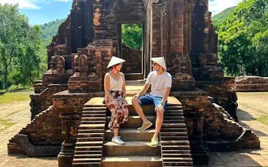 Visitors sitting on steps at Mỹ Sơn Sanctuary, Vietnam, with ancient temple ruins in the background.