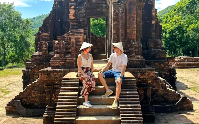 Visitors sitting on steps at Mỹ Sơn Sanctuary, Vietnam, with ancient temple ruins in the background.