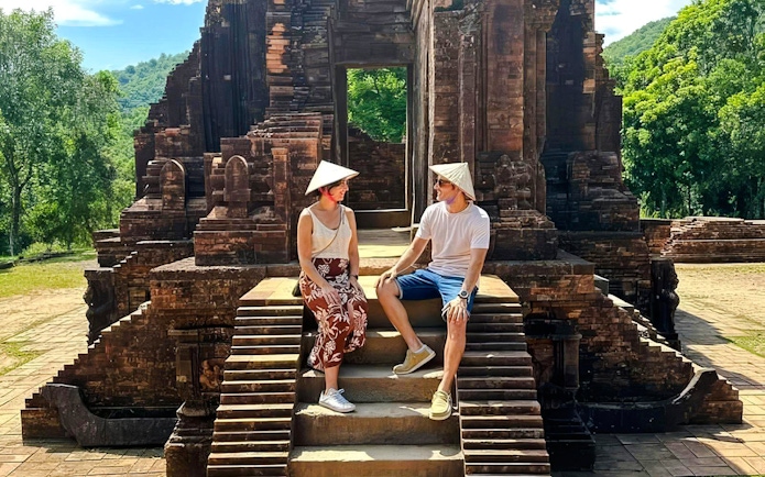 Visitors sitting on steps at Mỹ Sơn Sanctuary, Vietnam, with ancient temple ruins in the background.