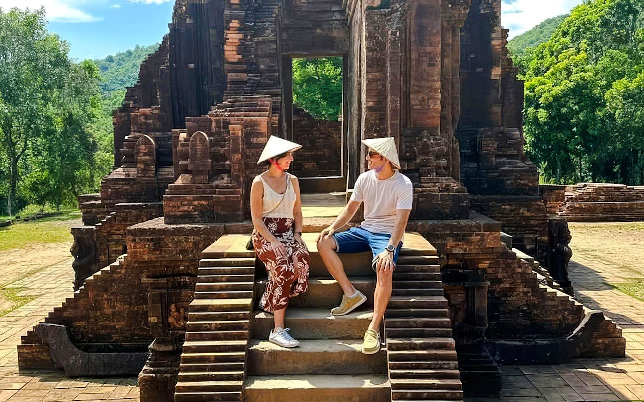 Visitors sitting on steps at Mỹ Sơn Sanctuary, Vietnam, with ancient temple ruins in the background.