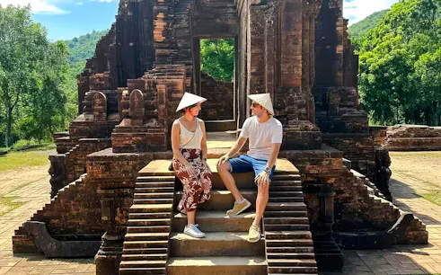 Visitors sitting on steps at Mỹ Sơn Sanctuary, Vietnam, with ancient temple ruins in the background.