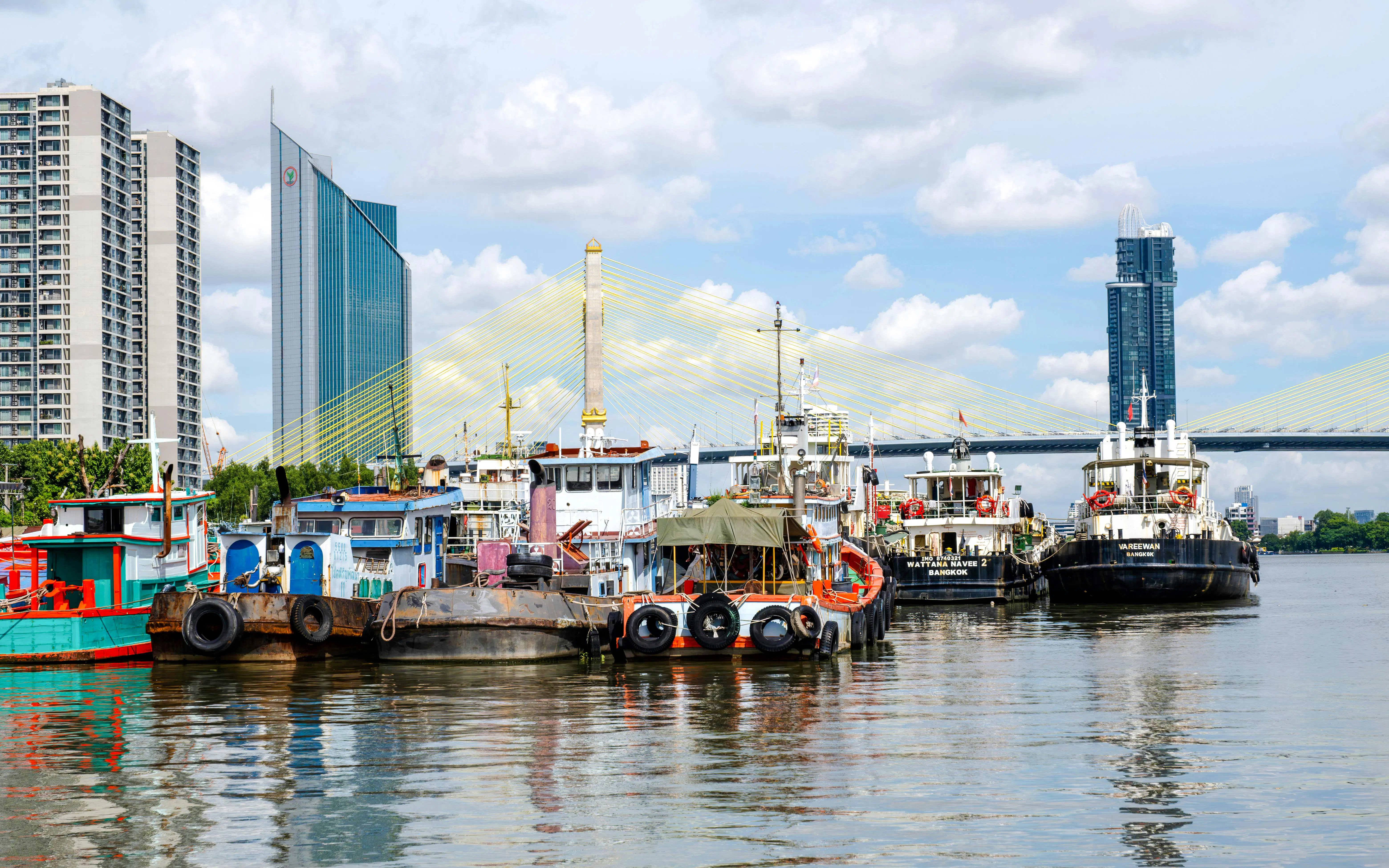Boats docked on the Chao Phraya River with Bangkok skyline and bridge in the background.
