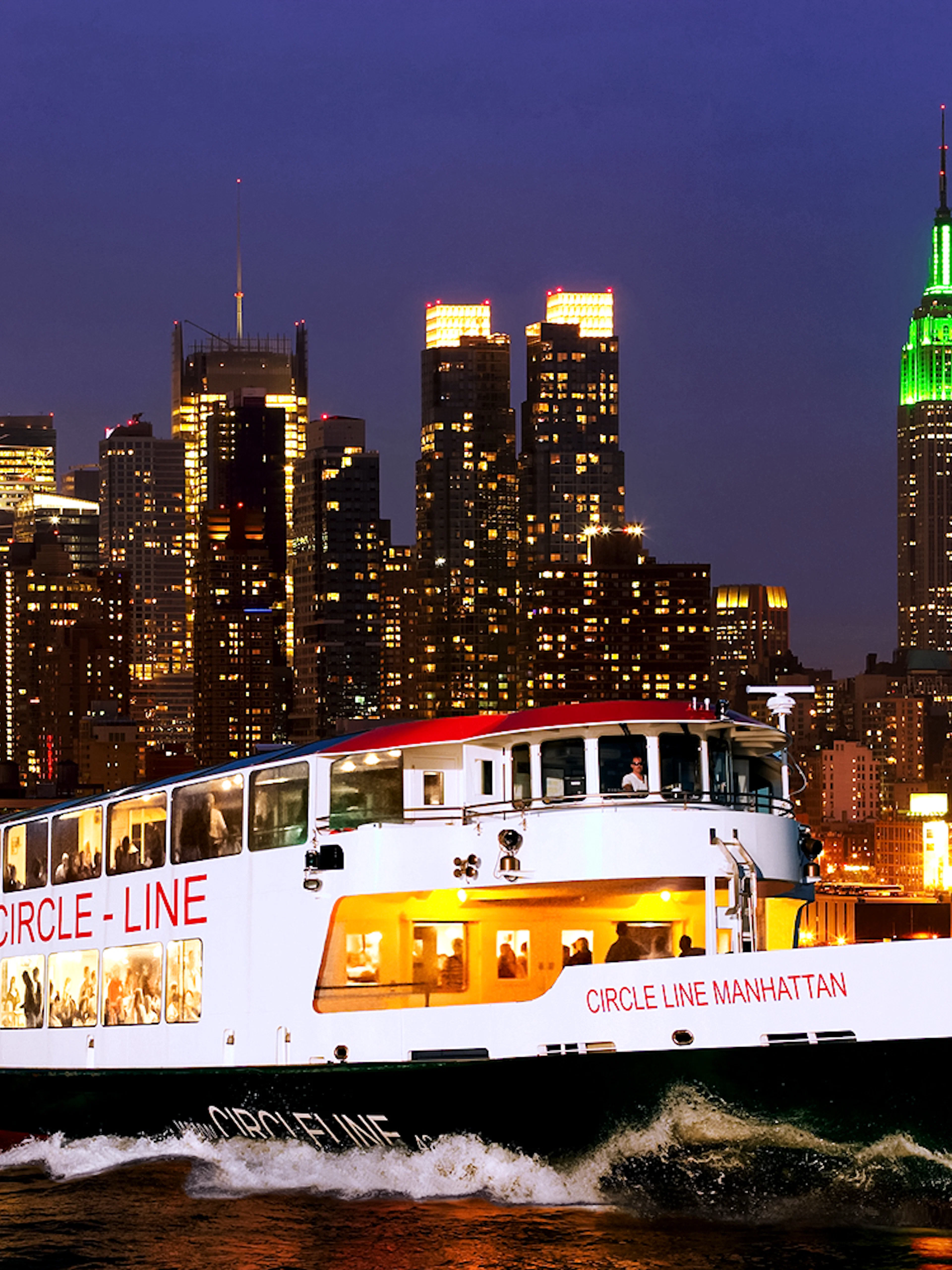 Tourists on Circle Line cruise with New York skyline illuminated at dusk.