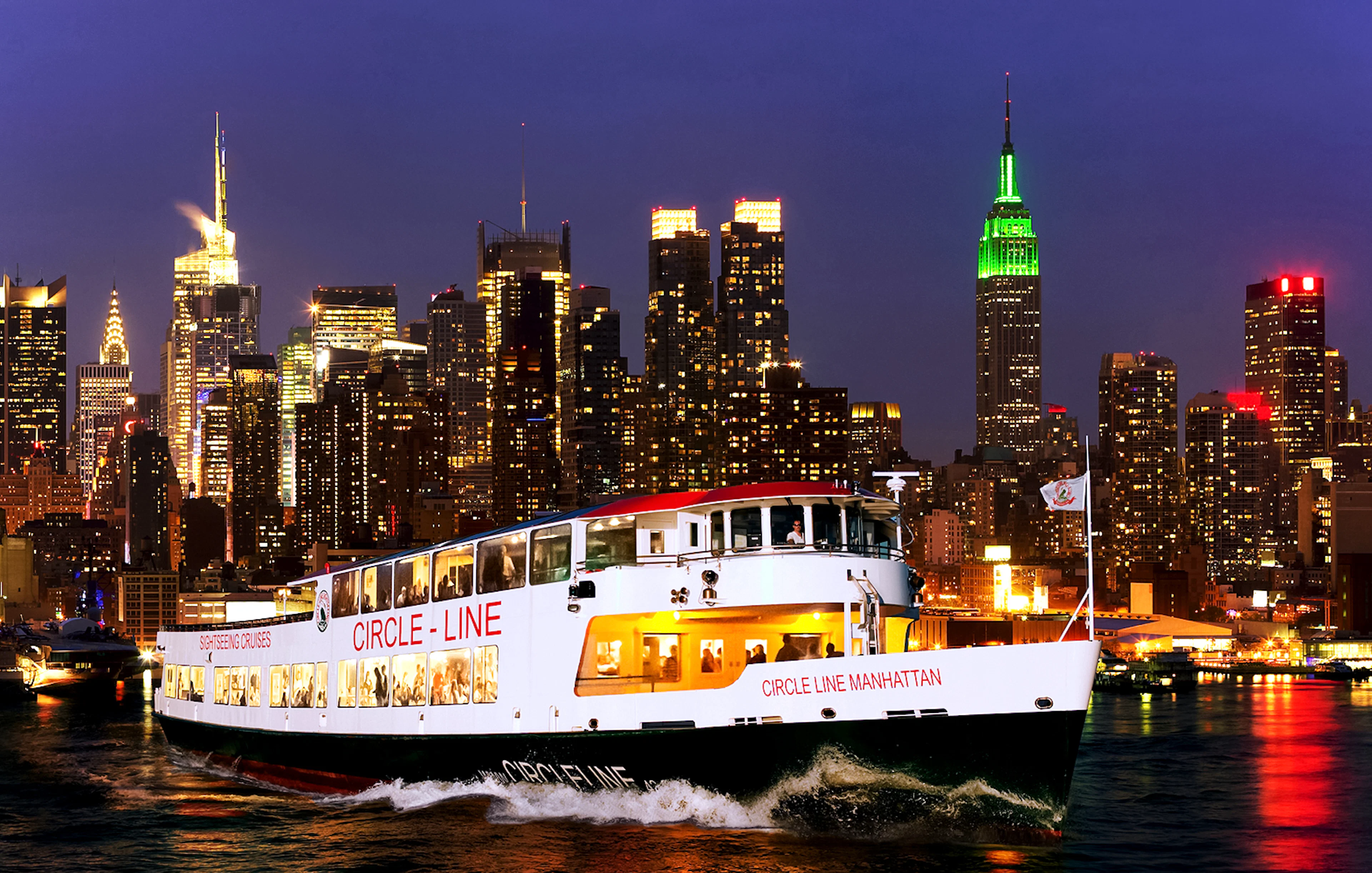 Tourists on Circle Line cruise with New York skyline illuminated at dusk.