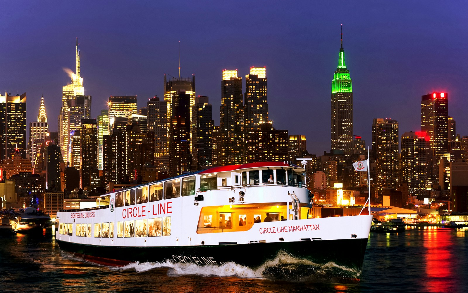 Tourists on Circle Line cruise with New York skyline illuminated at dusk.
