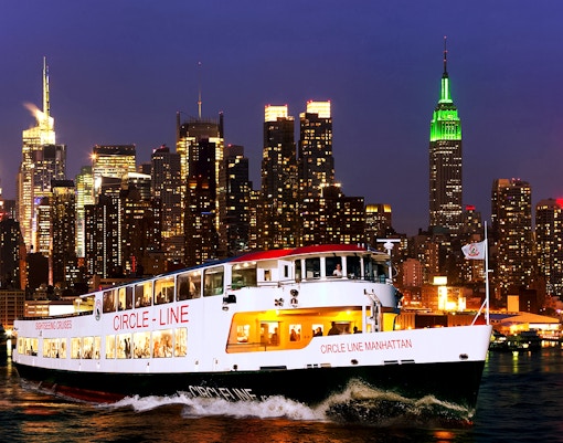 Tourists on Circle Line cruise with New York skyline illuminated at dusk.
