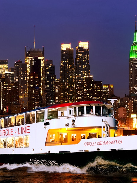 Tourists on Circle Line cruise with New York skyline illuminated at dusk.