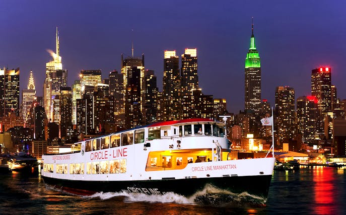 Tourists on Circle Line cruise with New York skyline illuminated at dusk.
