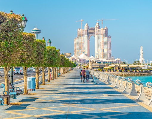 People walking along the Corniche promenade in Abu Dhabi with city skyline.