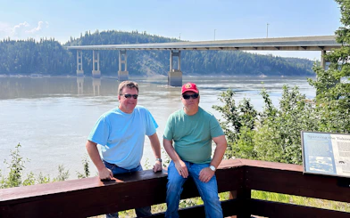 Two people at a scenic viewpoint by a river with a bridge in the background, Northern Lights & Arctic Circle Tour.