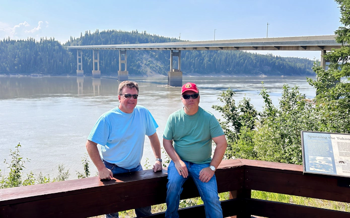 Two people at a scenic viewpoint by a river with a bridge in the background, Northern Lights & Arctic Circle Tour.