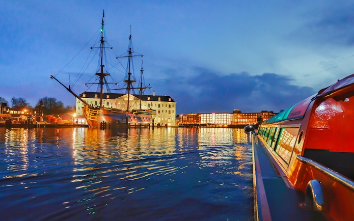 Amsterdam canal cruise at dusk with historic ship and illuminated buildings.
