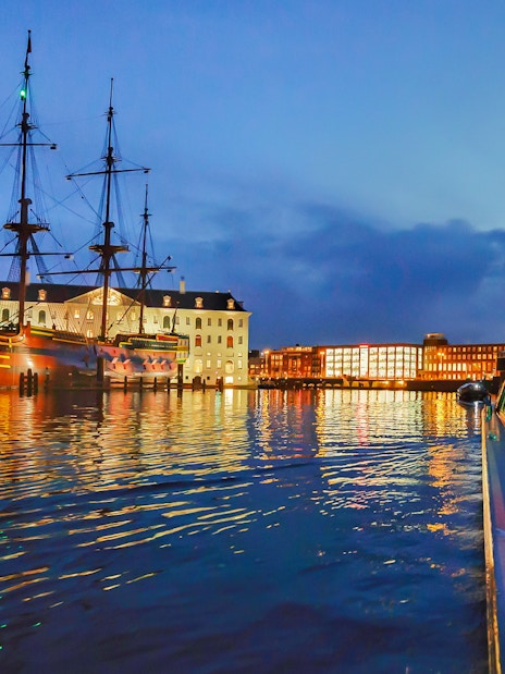 Amsterdam canal cruise at dusk with historic ship and illuminated buildings.
