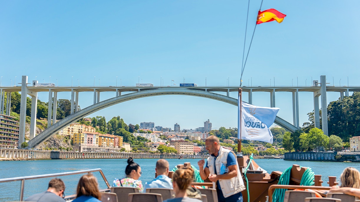 Tour guide explaining to tourists on Douro River cruise with Arrábida Bridge in Porto.