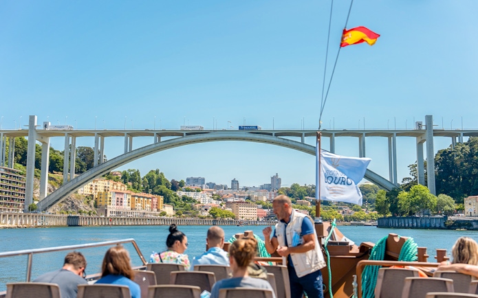 Tour guide explaining to tourists on Douro River cruise with Arrábida Bridge in Porto.
