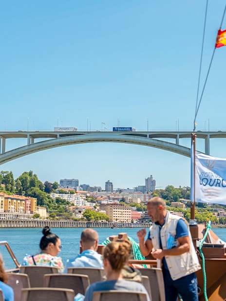 Tour guide explaining to tourists on Douro River cruise with Arrábida Bridge in Porto.