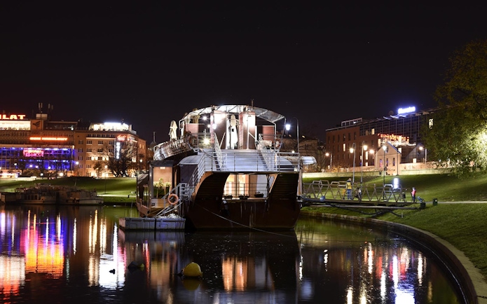 Night view of a boat docked on the Vistula River with city lights reflecting on the water.
