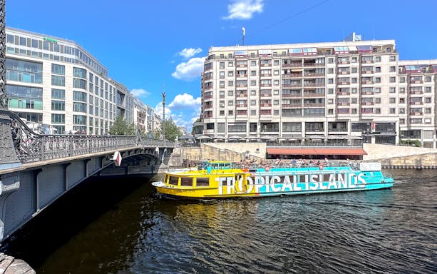 Berlin Spree River cruise boat passing under a bridge with city buildings in the background.