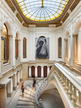 Grand staircase in a historic building with a large art piece under a stained glass dome.