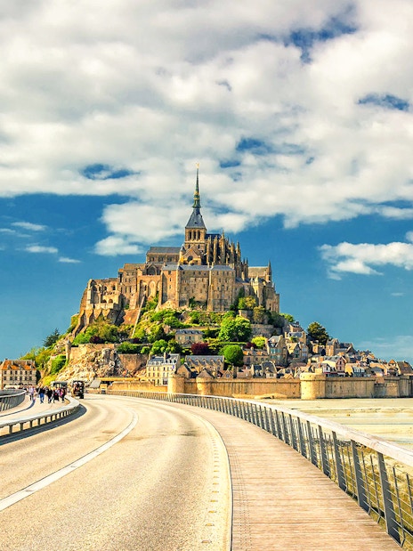 Mont Saint-Michel with bridge and visitors, France, under a blue sky.