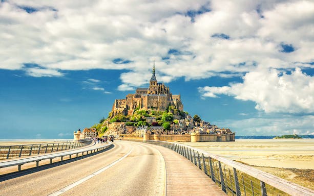 Mont Saint-Michel with bridge and visitors, France, under a blue sky.