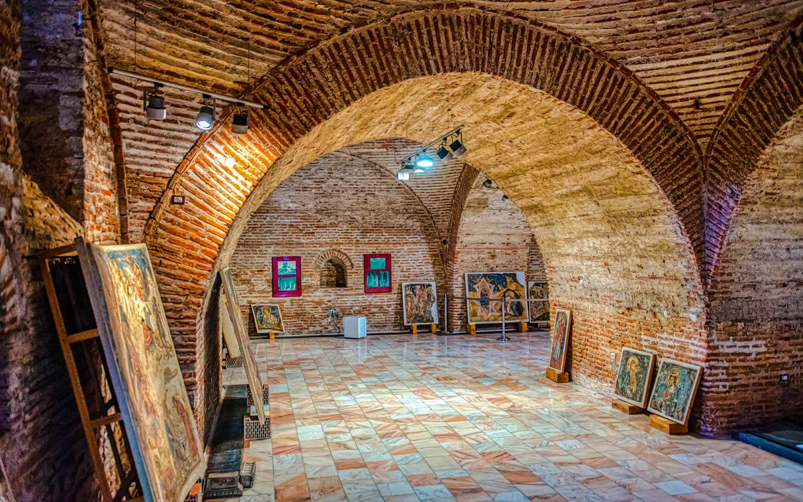Art gallery inside Mogosoaia Palace, Romania, featuring brick arches and paintings on display.