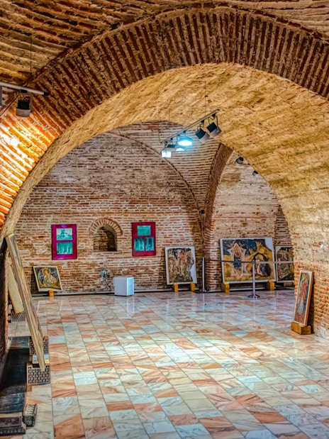 Art gallery inside Mogosoaia Palace, Romania, featuring brick arches and paintings on display.