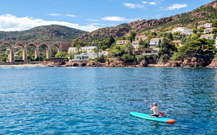 Paddleboarder on Villefranche bay with coastal homes and viaduct in background.