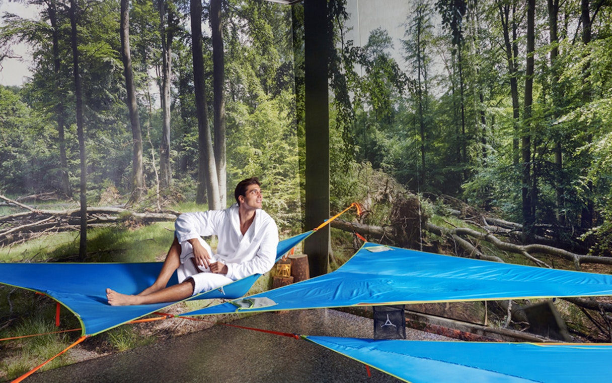 Man relaxing on a blue hammock at QC Terme Pré Saint Didier spa with forest backdrop.