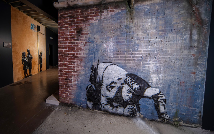 Street art on brick wall at Banksy Museum, Amsterdam, featuring a crouching figure.