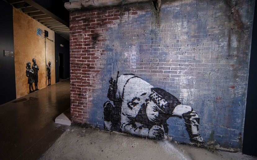 Street art on brick wall at Banksy Museum, Amsterdam, featuring a crouching figure.