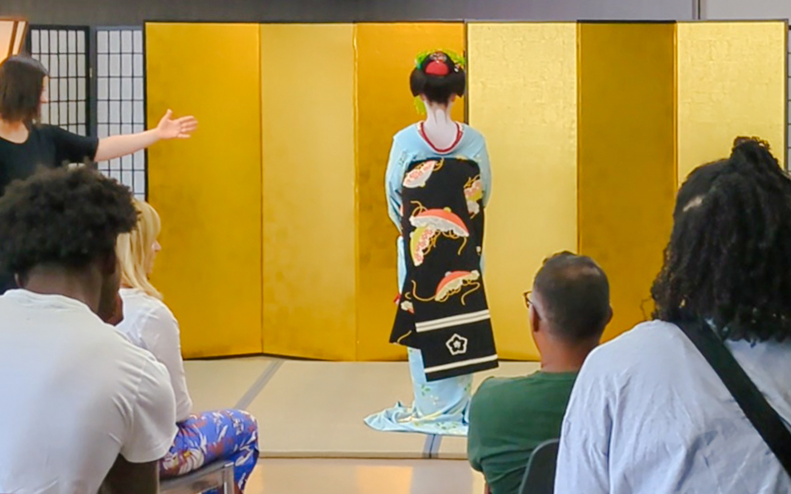 Maiko in light blue kimono with floral obi stands before an audience in Japan.