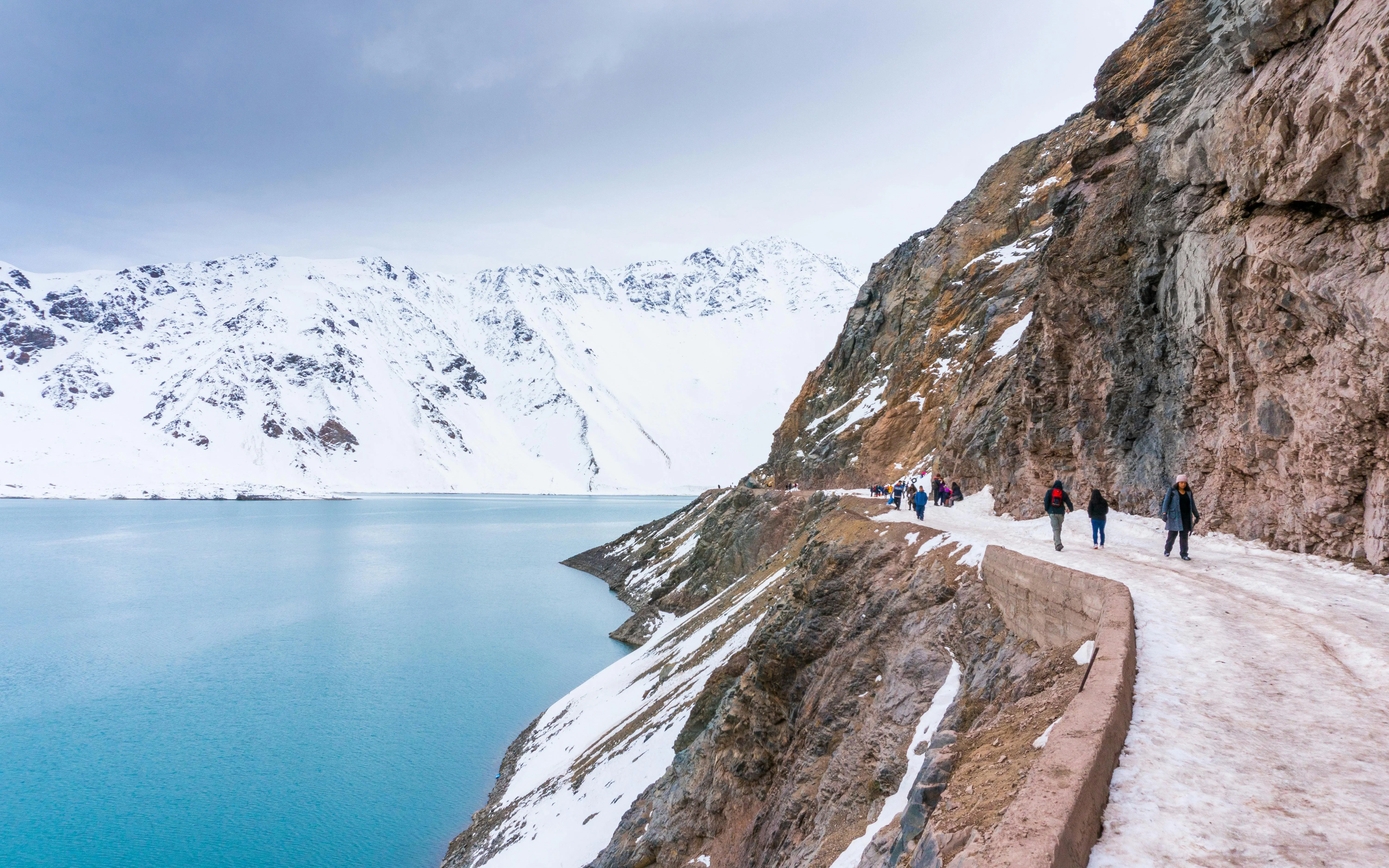 Hikers on a snowy trail along a turquoise lake at Cajon del Maipo, Chile.
