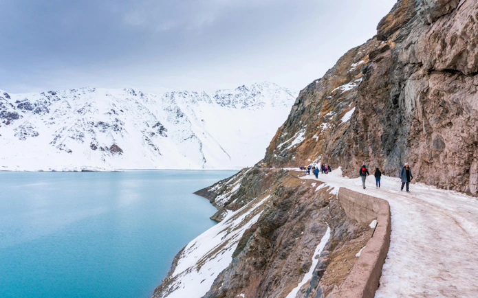 Hikers on a snowy trail along a turquoise lake at Cajon del Maipo, Chile.