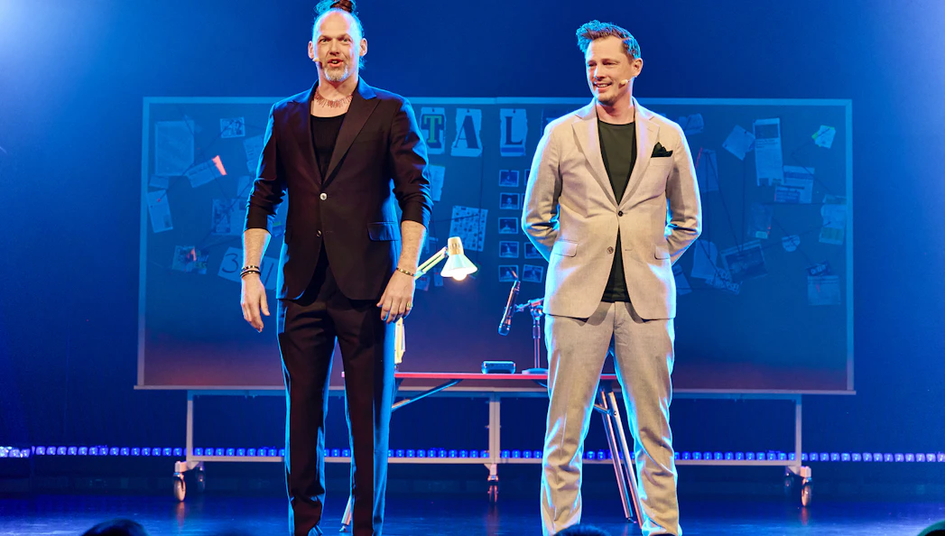 Two performers on stage with a board of notes and photos in the background.