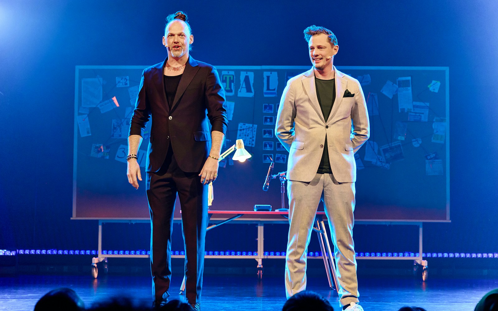 Two performers on stage with a board of notes and photos in the background.