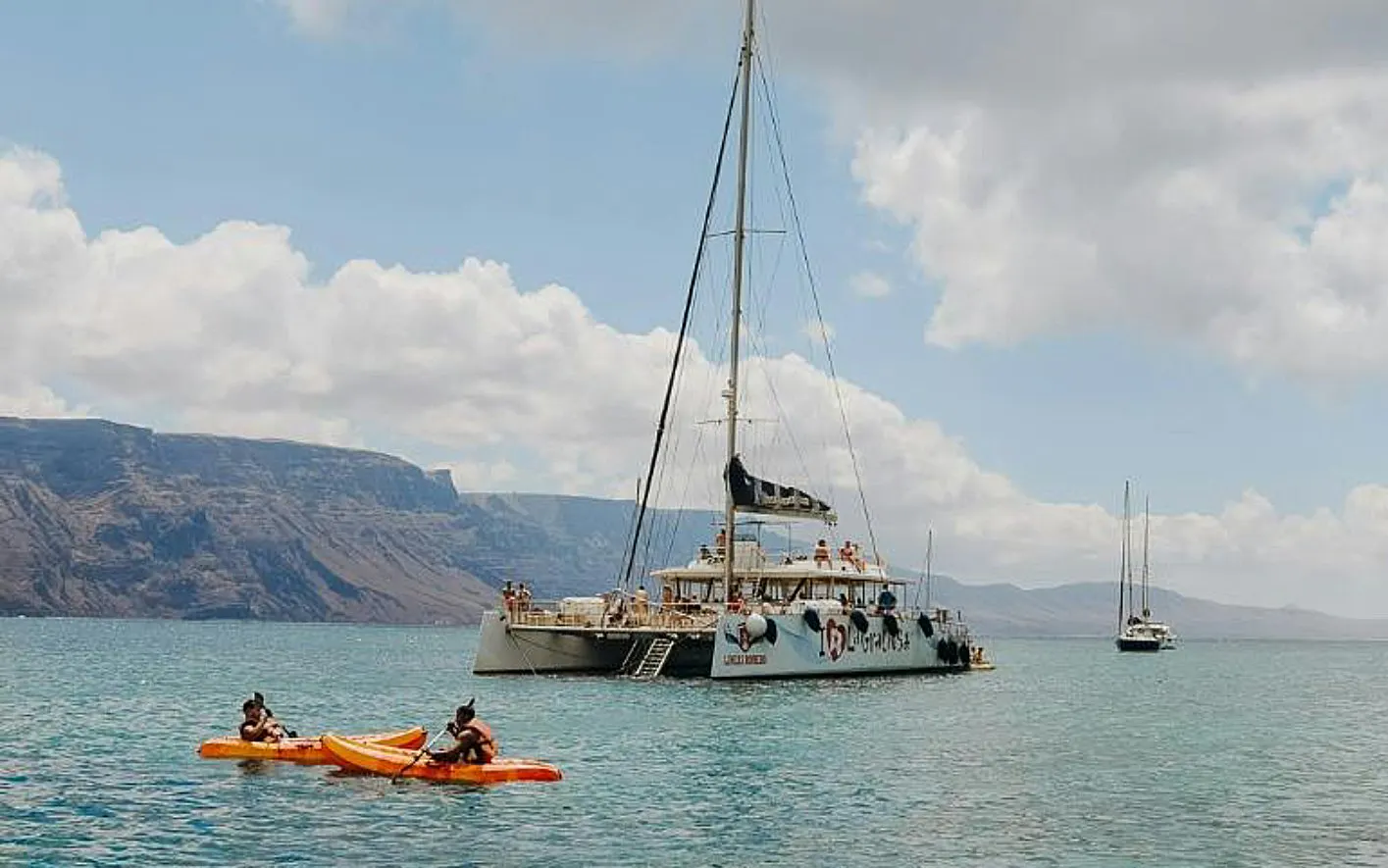 Catamaran sailboat cruising near La Graciosa Island, Lanzarote, with kayakers in foreground.